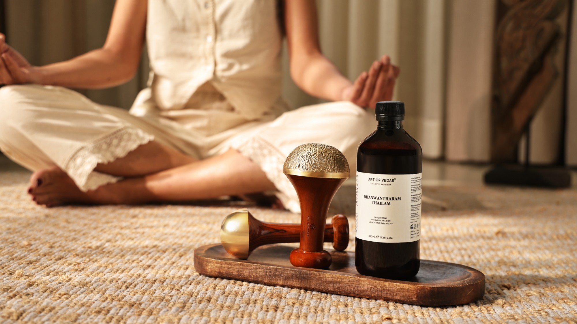 Two Art of Vedas Kansa wands and a bottle of Dhanwantharam Thailam on a wooden tray, a woman seated in meditation behind